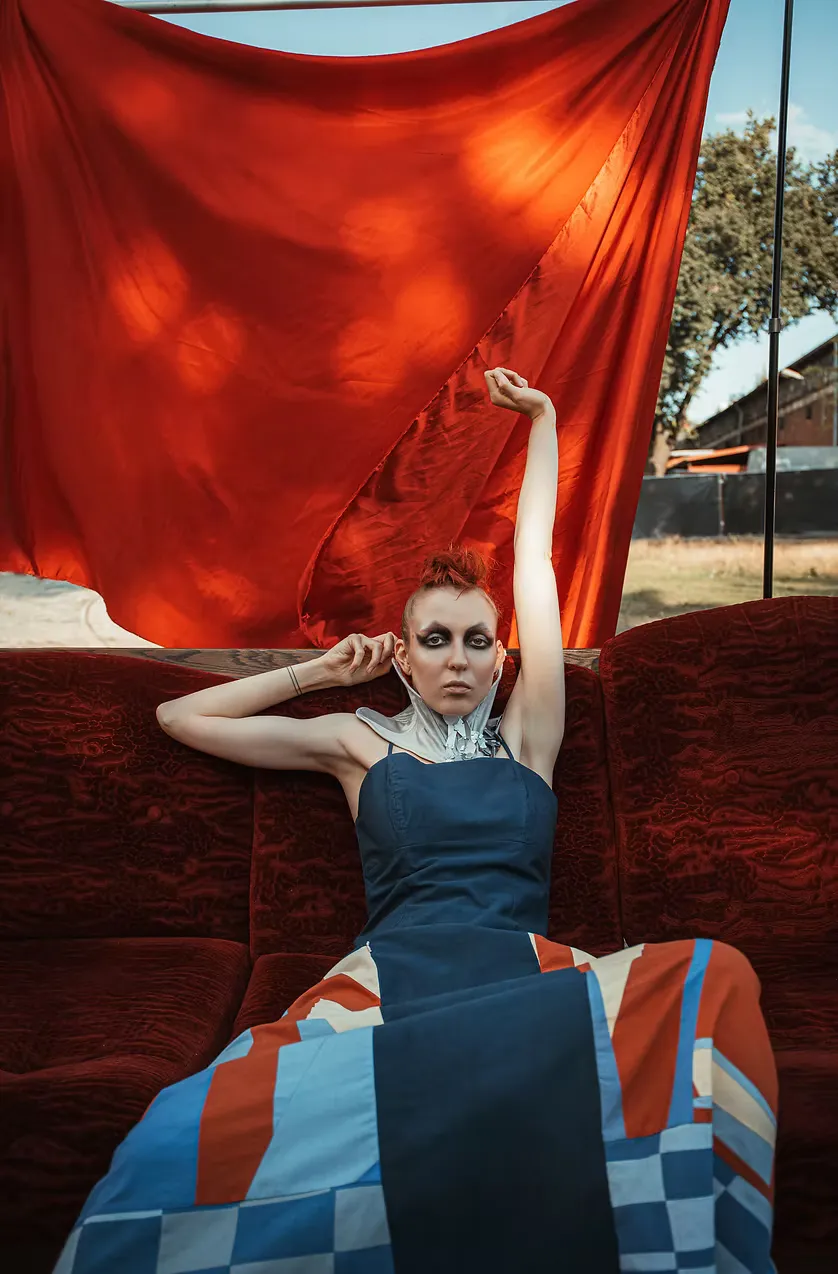A model with platinum hair and silver eye makeup reclines on the red velvet sofa, one arm raised holding red fabric. A patchwork geometric skirt and slate dress cut a bold silhouette against open sky.