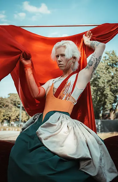 A model with platinum hair and tattoos raises billowing red fabric against a blue sky, wearing the teal skirt and orange bodice from the original collection. Costume as gesture — defiant and celebratory.