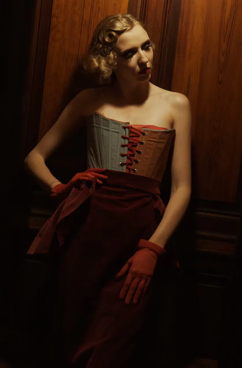 A figure with vintage-waved hair leans against dark paneled wood wearing a structured gray corset with bold red lacing and matching red gloves. The intimate garment made outerwear — craftsmanship as statement, history worn on the surface.