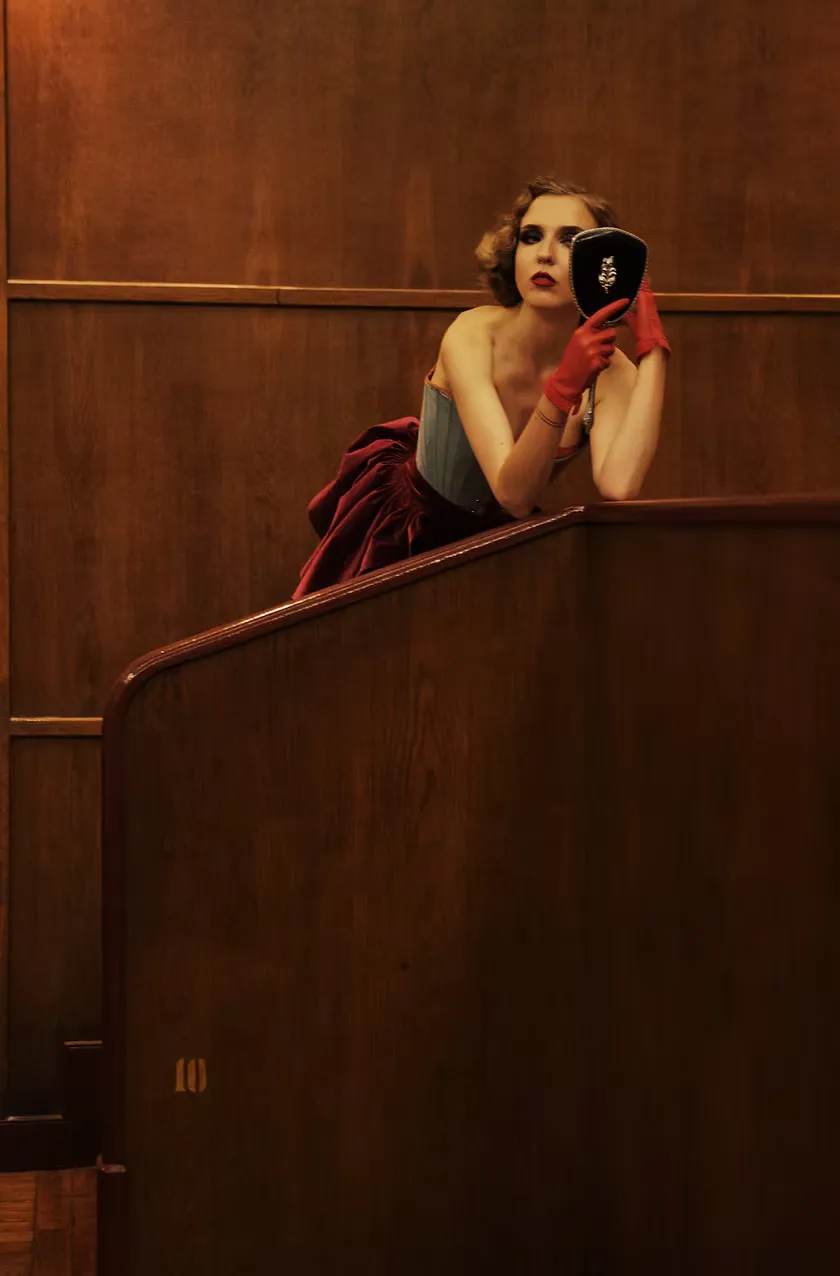Framed by a theater box railing, a figure in a gray strapless top and red cape leans forward holding a hand mirror. Bold red gloves and a vivid lip complete the tableau — the Other as audience and spectacle simultaneously.