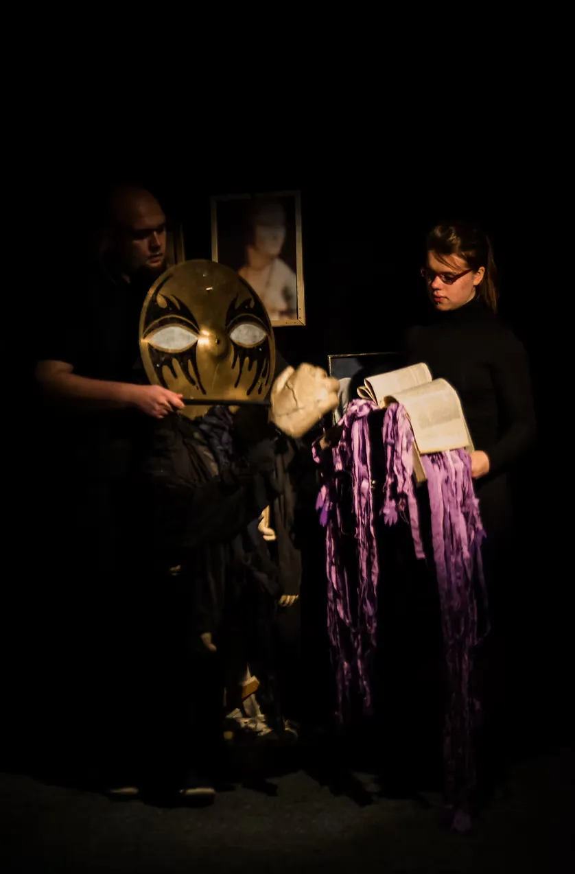 Two performers on a dimly lit stage — one holds aloft a large ornate gold mask with bold eye markings, the other cradles cascading purple fringe. A framed image hangs in the background.