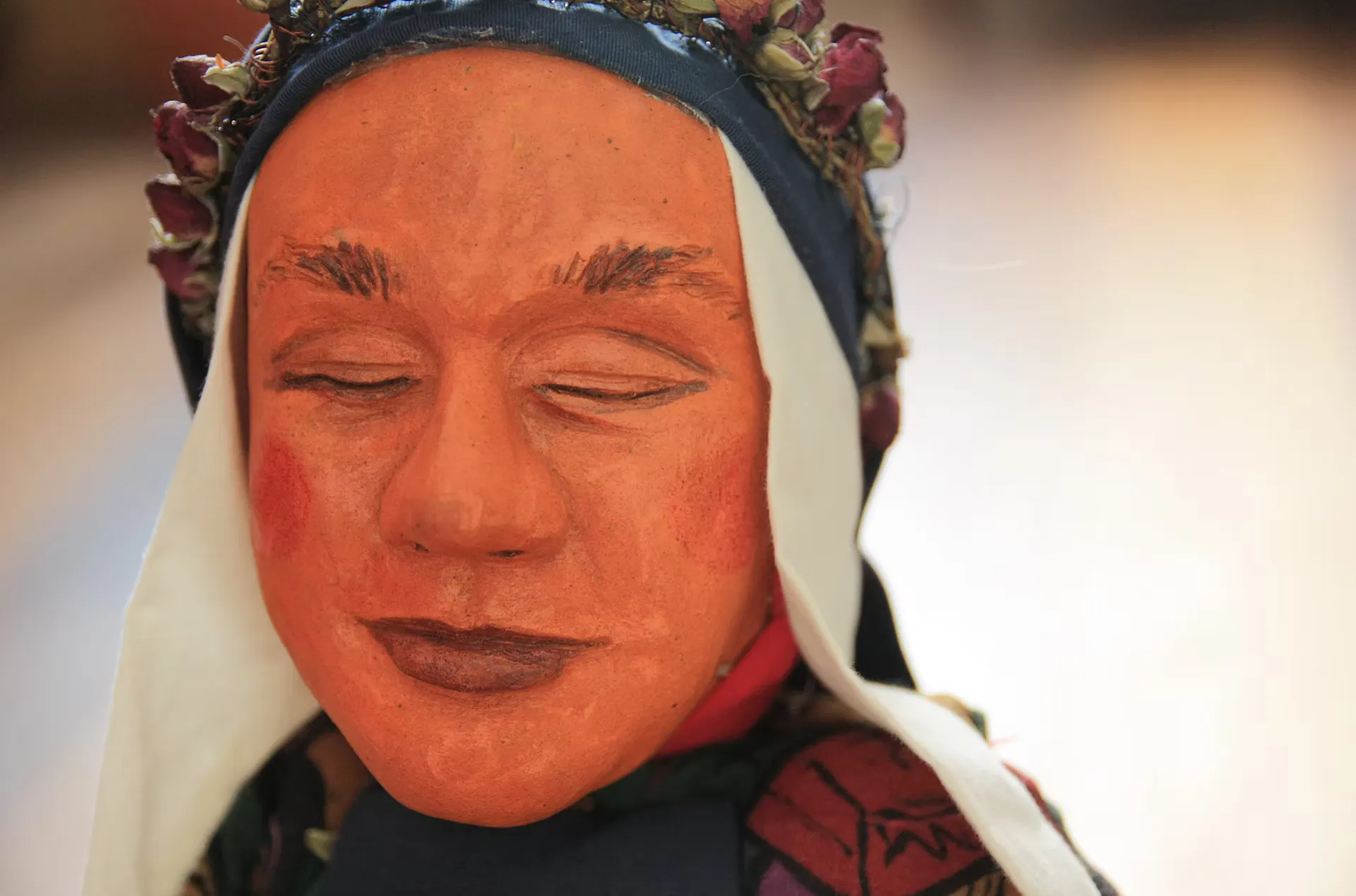 Portrait of a puppet head with a terracotta-painted face, closed eyes, and rosy cheeks, wearing a white cloth headdress with dark blue trim and a crown of dried fabric roses. A folk character rendered with tenderness and craft.