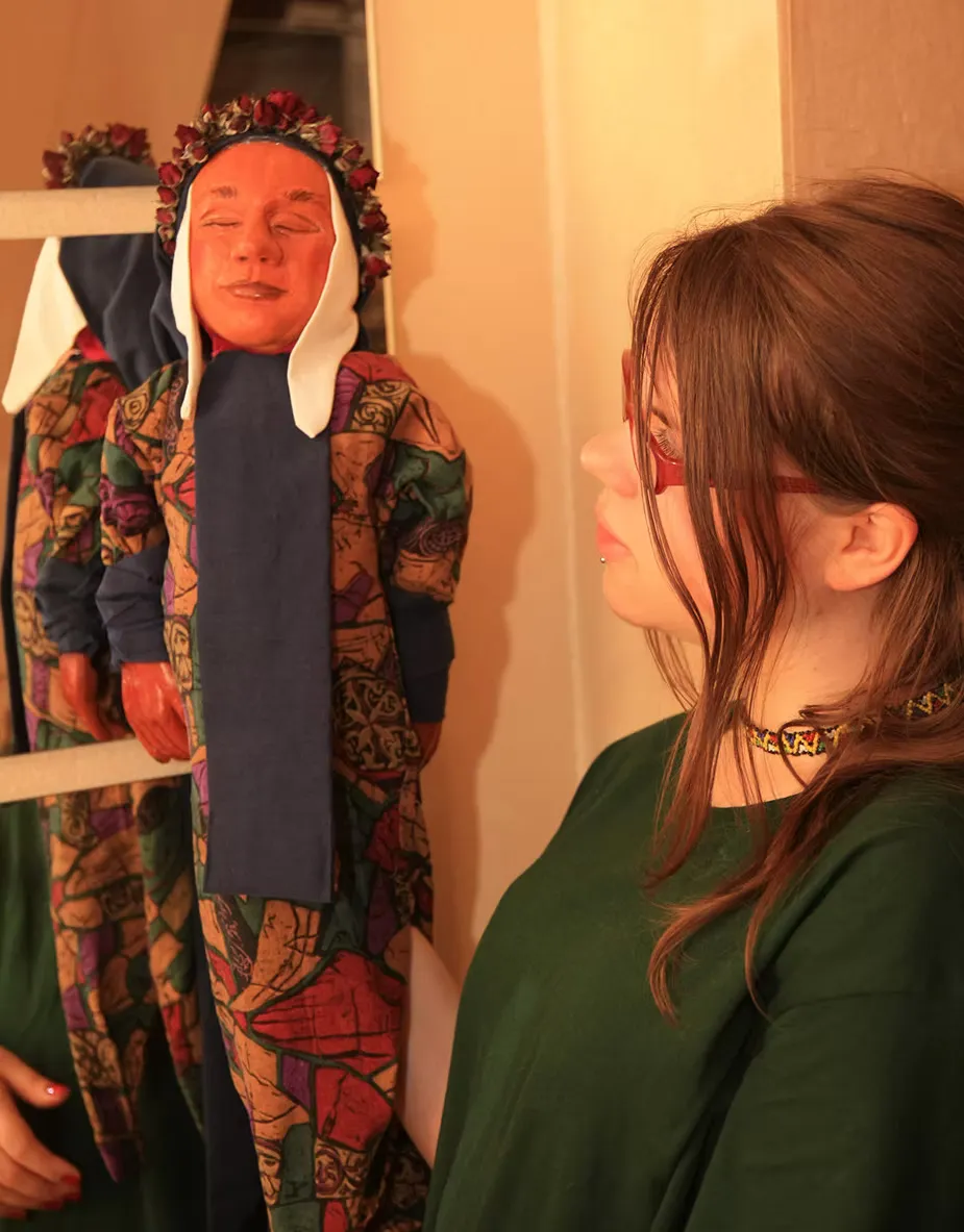 The artist looks quietly at a puppet hanging from a wooden rack — the same terracotta folk figure in colourful patterned clothing. An intimate moment in the rehearsal space.