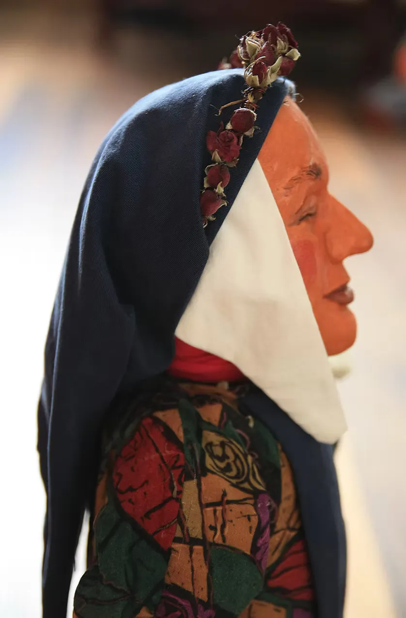 Profile view of the terracotta folk puppet head — the painted orange face in sharp side relief, white and dark blue headdress adorned with dried fabric roses. Handcraft foregrounded in detail.