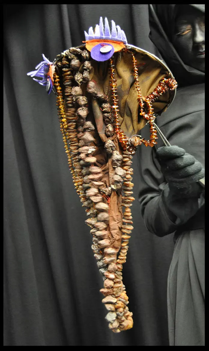 A tall totemic staff held by a black-gloved hand — organic forms of shells, seeds, and bone-like shapes strung with amber beads, crowned with fish-like silhouettes and a violet accent. Ritualistic and strange.