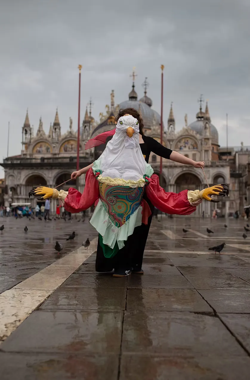 A performer in a large handmade seagull costume stands arms-wide in Venice's Piazza San Marco on a rain-slicked day, St. Mark's Basilica behind them, pigeons scattered across the stones. Costume as public spectacle.