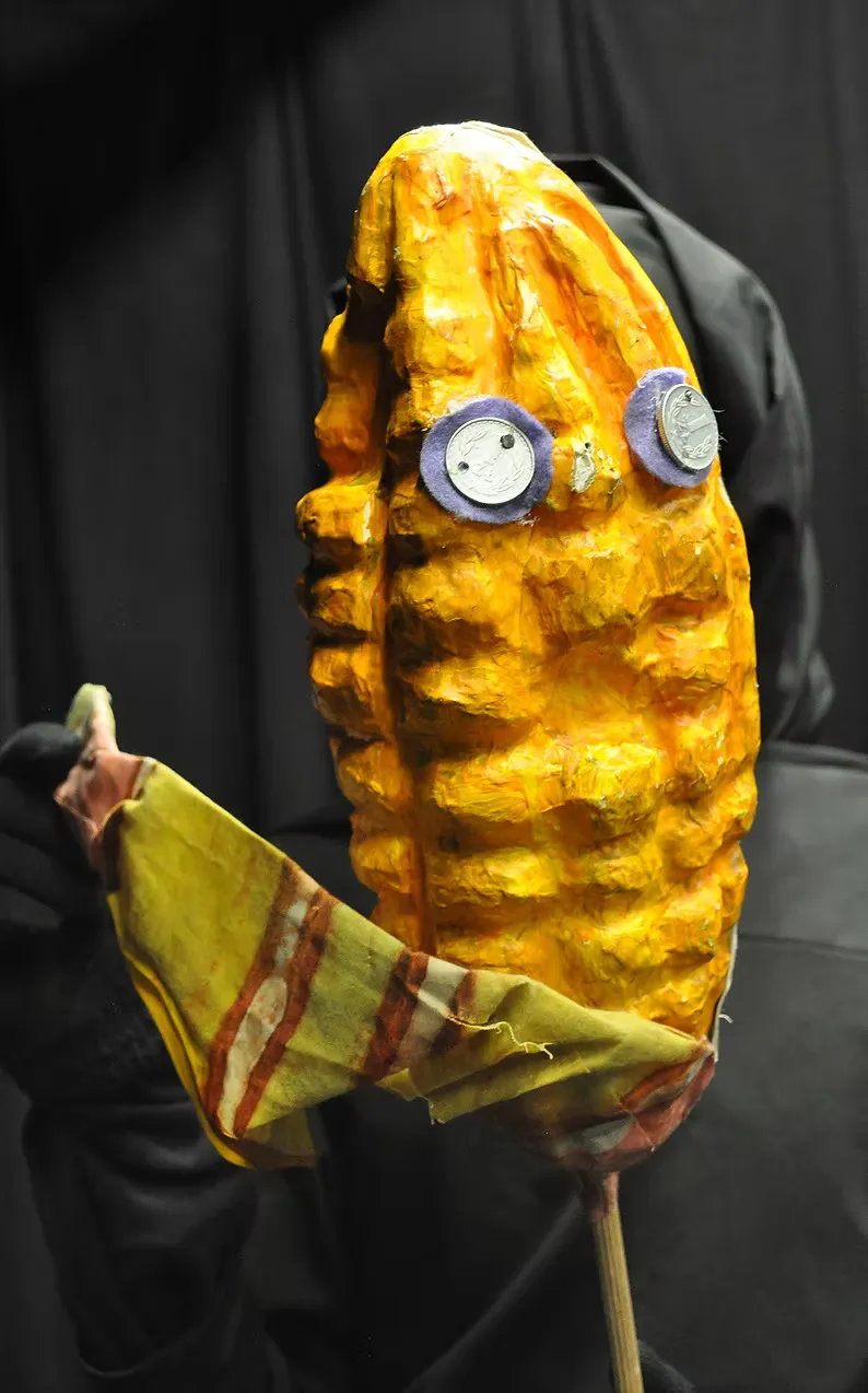 A large papier-mâché puppet head in the form of a stylised fish — ridged orange-yellow surface, button eyes with hand-drawn smiley faces, a green leaf-like fin. Held on a stick against darkness. Playful and strange.