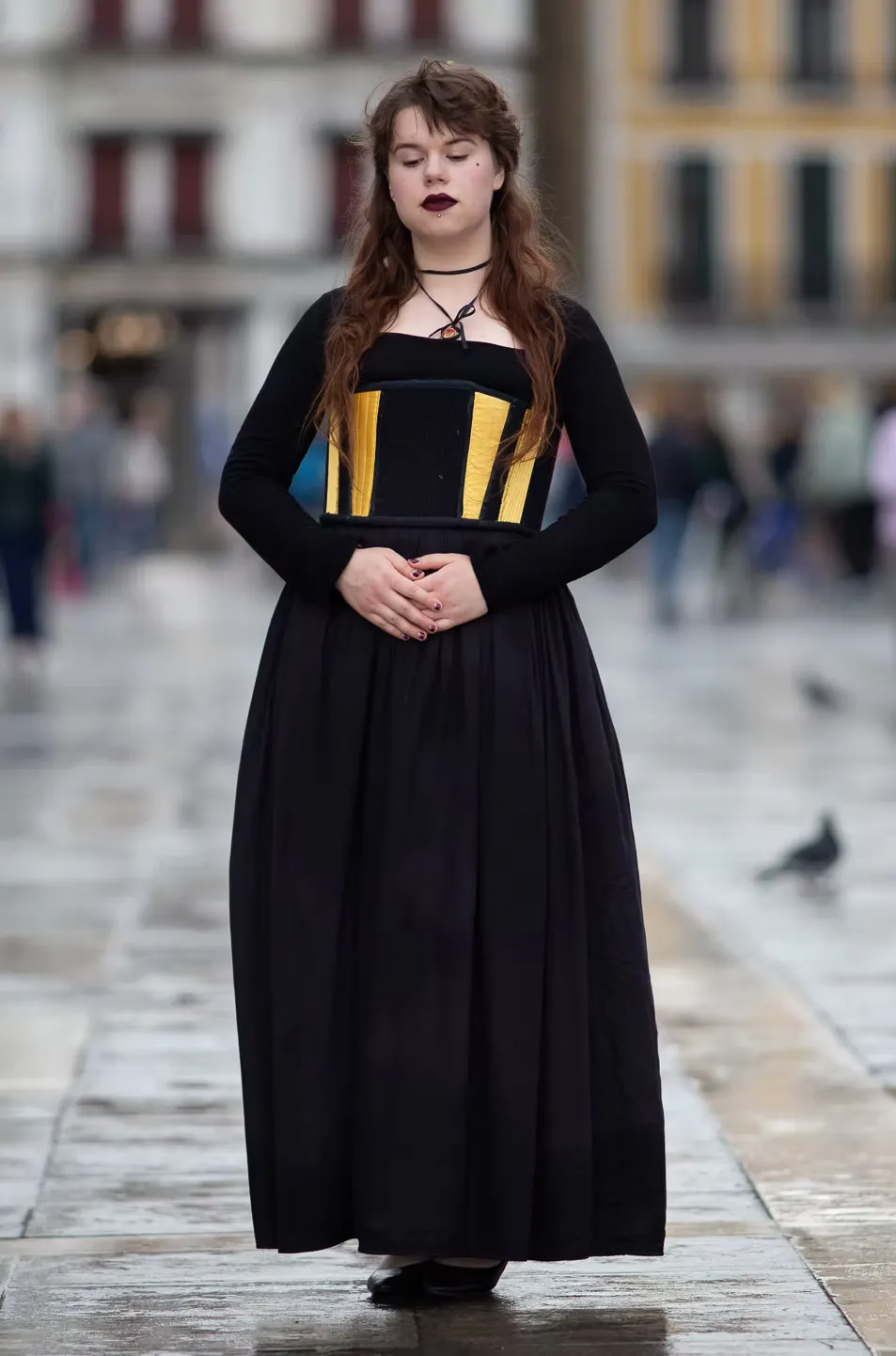 The artist stands in a public square wearing a long black dress with a structured bodice of yellow, blue, and black panels with red lacing. Dark hair and lips. A costume brought out of the theatre and into the city.