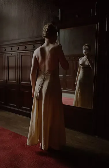 A figure in a backless floor-length gold satin gown stands before a large ornate mirror in a dim wood-paneled room. Her reflection gazes outward. A meditation on the threshold between private self and public persona.